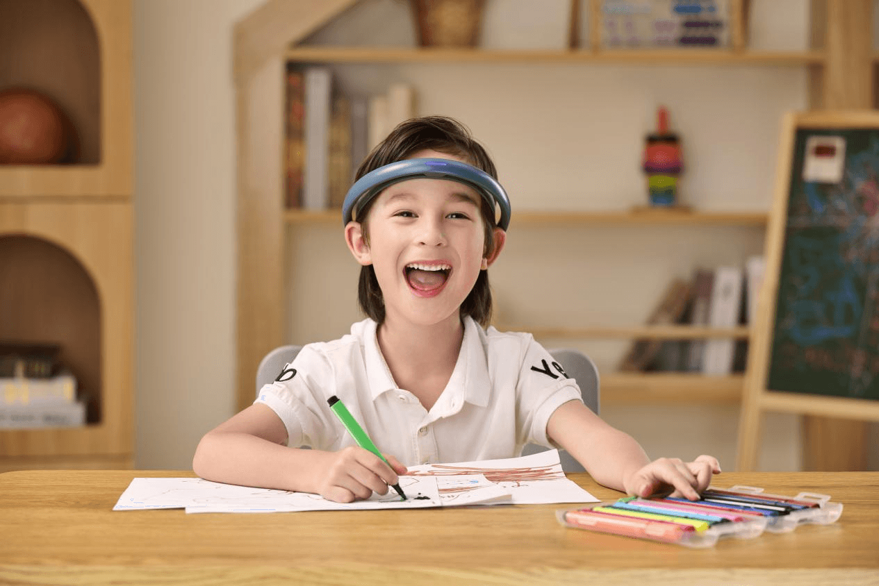 A happy young boy wearing the BrainCo FocusXin headband smiles while drawing at his desk, showing the positive engagement from the training.