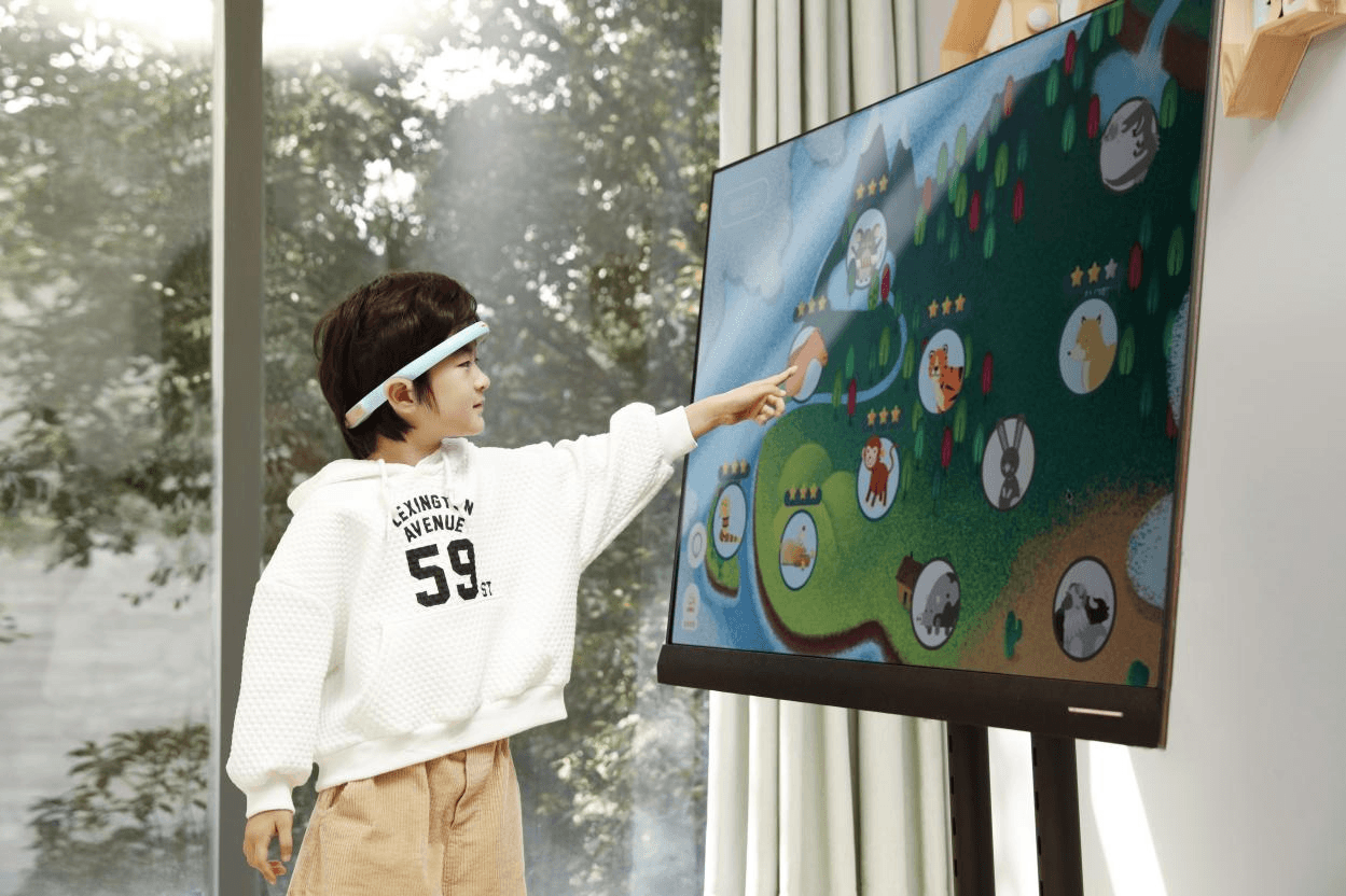 A young child wearing a BrainCo BCI headband pointing at a large screen, engaging with the StarKids gamified training system.