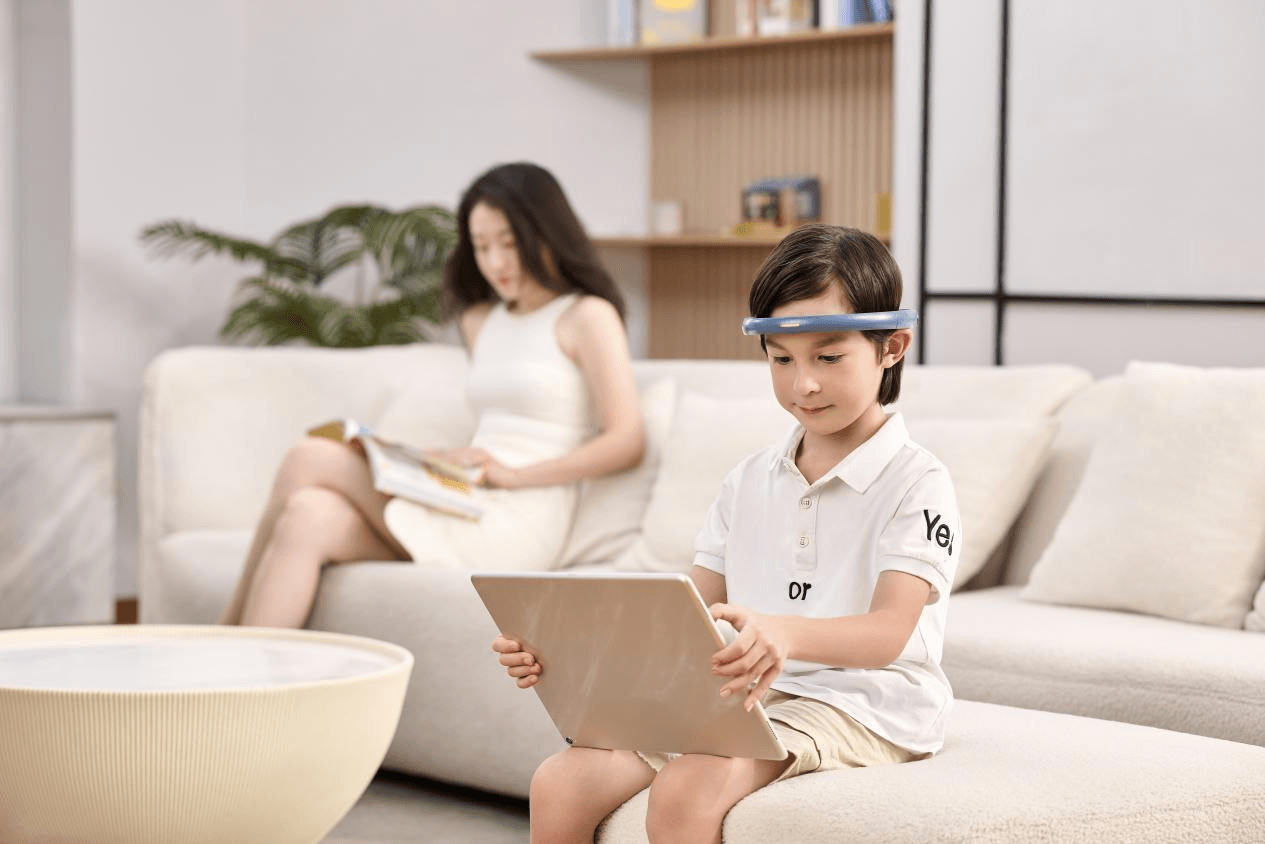 一个男孩在客厅沙发上佩戴着BrainCo FocusXin专注力训练头环,正使用平板电脑进行互动训练,母亲在后方阅读,展现了和谐的家庭科技应用场景。